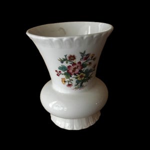 May include: A white ceramic vase with a flared top and a bulbous midsection. The vase features a floral design with red, pink, yellow, and green flowers. The base has a scalloped edge, and the background is black.