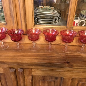 May include: Six red glass stemmed goblets with etched designs. The goblets are arranged in a row on a wooden surface.