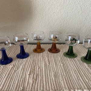 May include: Six stemmed wine glasses with different colored bases. The glasses are decorated with gold lettering and designs. The glasses are arranged in a row on a white textured surface.