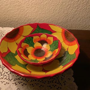 May include: Two hand-painted ceramic bowls, one nested inside the other. The bowls feature a sunflower design with yellow petals, green leaves, and a red rim. The larger bowl is approximately 12 inches in diameter, and the smaller bowl is about 6 inches.