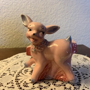 May include: A vintage pink ceramic deer figurine with a winking expression. The deer has a floral collar and a small planter on its back. The figurine is painted with shades of pink, blue, and red.