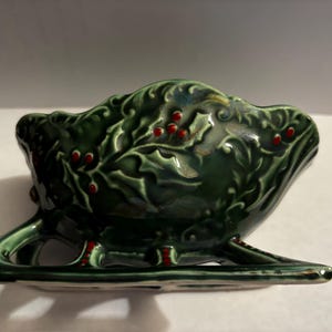 May include: A green ceramic bowl shaped like a sleigh with holly and red berries. The bowl is glazed with a green and iridescent finish.