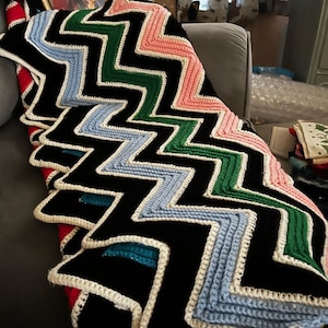 May include: A handmade, colorful crochet blanket with a bold zigzag pattern. The blanket features black, white, blue, green, and pink sections. The edges are finished with a white border, creating a striking visual contrast. This cozy blanket is perfect for adding a pop of color to any room.