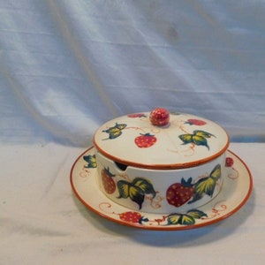 May include: A white ceramic butter dish with a lid and a matching plate. The dish is decorated with a strawberry and leaf design in red, green, and yellow.