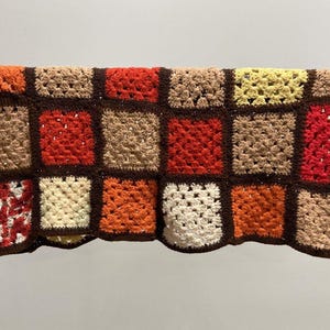 May include: A handmade crochet blanket with a patchwork design. The blanket features squares in shades of red, orange, beige, and yellow, bordered by dark brown. The blanket is displayed against a neutral background.
