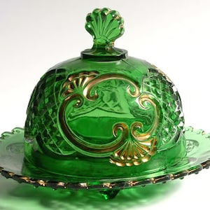 May include: A green glass butter dish with a gold-painted design. The dish has a domed lid with a scalloped edge and a small finial on top. The base of the dish is a scalloped plate.