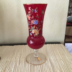 May include: A red glass vase with a gold rim and floral design. The vase has a round base and a tall, slender neck. The flowers are painted in shades of red, blue, yellow, and white.