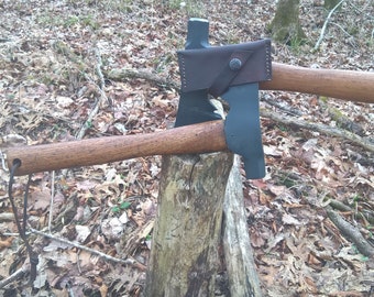 Peut inclure: Deux haches avec des manches en bois et des lames gris foncé sont présentées sur une souche d'arbre. Un étui en cuir marron protège la lame d'une hache. Les haches sont à l'extérieur, entourées de feuilles et de branches.