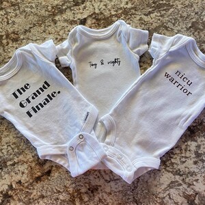 May include: Three white baby onesies with black text. The onesies say "The Grand Finale", "Tiny & Mighty", and "Nicu Warrior".