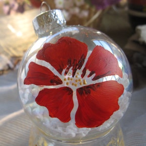 May include: A clear glass Christmas ornament with a hand-painted red flower design. The flower has a white center with gold accents.