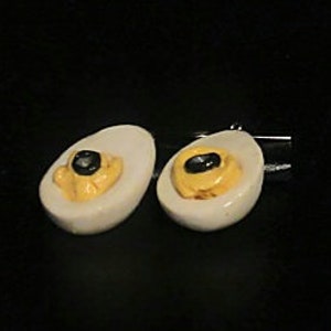 May include: A pair of white ceramic cufflinks shaped like halved boiled eggs with yellow yolks and black peppercorns.