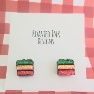 May include: A pair of small, colorful, layered cake earrings. The earrings are made of polymer clay and are shaped like a slice of cake. The cake is green, yellow, and pink, and is topped with a layer of chocolate. The earrings are on a white card with the text "Roasted Ink Designs".