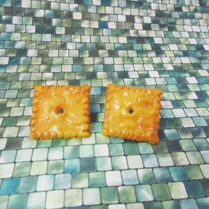 May include: A pair of yellow, square-shaped earrings that resemble a popular cracker brand. The earrings have a hole in the center and a textured edge.