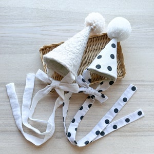 Puede incluir: Dos gorros de fiesta blancos con diferentes patrones, uno con un pompón blanco y el otro con lunares negros. Ambos gorros tienen lazos de cinta blanca.