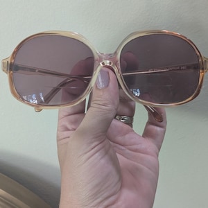 May include: Oversized, light brown sunglasses with dark lenses. The glasses have a rounded shape and a thin, gold-colored frame. The text "AO American Optical 140" is visible on the arm of the glasses.