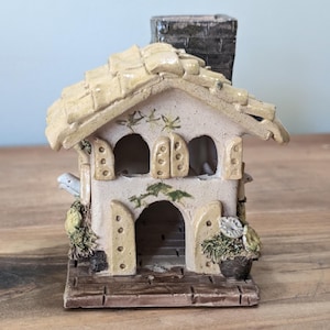 May include: A ceramic miniature house with a beige roof and walls, featuring arched windows and a chimney. Decorative elements include small plants and a flower pot, with a weathered, rustic appearance. The house sits on a wooden base.