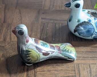 Könnte beinhalten: Zwei handbemalte Keramik-Vogelfiguren. Ein Vogel ist weiß mit blauen und braunen Blumendesigns. Der andere Vogel ist braun, grün und gelb mit einem Blumendesign. Beide Vögel haben schwarze Augen.