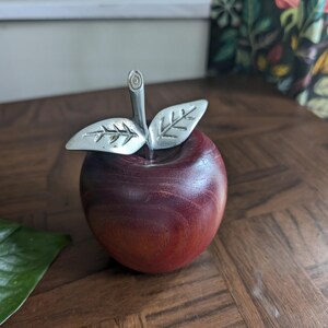 K&ouml;nnte beinhalten: Eine dekorative Apfelskulptur mit einem tiefroten Holzk&ouml;rper und silberfarbenen Metallbl&auml;ttern und -stiel. Der Apfel ist ein solides, poliertes St&uuml;ck, wahrscheinlich f&uuml;r die Inneneinrichtung oder als Briefbeschwerer.