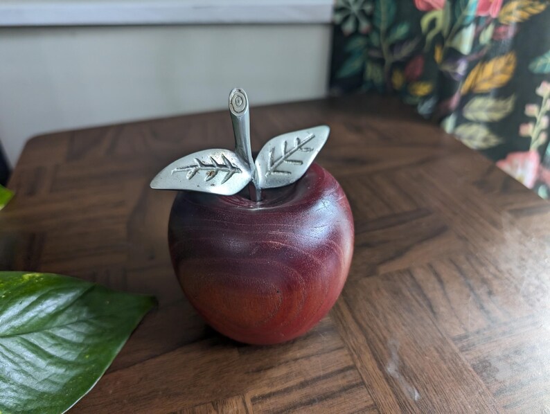 K&ouml;nnte beinhalten: Eine dekorative Apfelskulptur mit einem tiefroten Holzk&ouml;rper und silberfarbenen Metallbl&auml;ttern und -stiel. Der Apfel ist ein solides, poliertes St&uuml;ck, wahrscheinlich f&uuml;r die Inneneinrichtung oder als Briefbeschwerer.