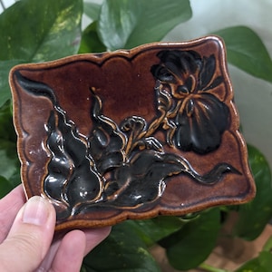 Puede incluir: Un pequeño plato rectangular de cerámica con un esmalte marrón y un diseño floral negro. El diseño presenta una flor grande con hojas y tallos.