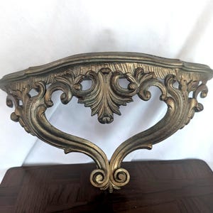 May include: Ornate, antique-style wall shelf in a gold and bronze finish. The shelf features intricate scrollwork and a curved, heart-shaped support. The shelf is designed to be mounted on a wall and is made of a durable material.