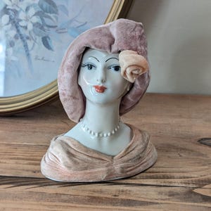 May include: A vintage porcelain bust of a woman with a pink velvet hat and matching draped bodice. The bust features a pearl necklace, red lips, and a peach-colored rose accent on the hat. The bust is on a wooden surface.