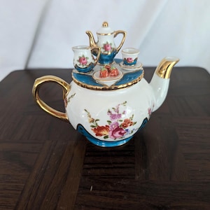 May include: A white porcelain miniature tea set with gold trim, featuring a teapot, two cups, and a small plate. The set is decorated with floral patterns in pink, red, and purple, with a blue border.