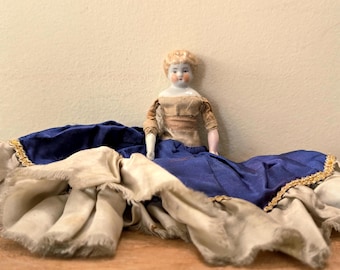 Könnte beinhalten: Eine Vintage-Porzellanpuppe mit blonden Haaren und einem bemalten Gesicht, die ein beiges und blaues Kleid trägt. Die Puppe liegt auf einem cremefarbenen Stoff mit einem blauen Rock, der mit Gold verziert ist.