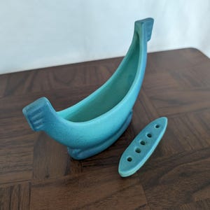 Op de afbeelding: Een mat, blauwgroen keramieken vaasje in de vorm van een gondel met een gebogen, langwerpig lichaam en verhoogde uiteinden. Naast het hoofdonderdeel ligt een bijpassend ovaal keramieken inzetstuk met vijf gaten voor het schikken van bloemen.