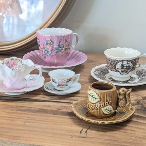May include: A collection of four teacups and saucers. The teacups are decorated with floral patterns and the saucers are white with gold trim. One teacup is brown with a squirrel handle and the text "Colorado" on the side.