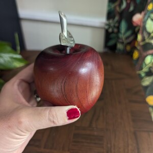 K&ouml;nnte beinhalten: Ein Holzapfel mit dunkelroter Oberfl&auml;che und einem silbernen Metallblatt und -stiel. Der Apfel wird in einer Hand gehalten. Der Hintergrund umfasst gr&uuml;ne Pflanzen und einen floral gemusterten Vorhang.