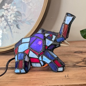 May include: A stained glass elephant lamp in a mosaic pattern. The elephant is composed of blue, red, and purple glass pieces. The lamp has a black cord and two white tusks. The lamp is on a wooden surface.