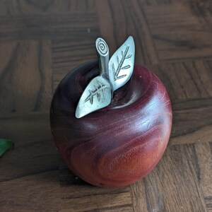 K&ouml;nnte beinhalten: Dekorative Apfelfigur aus rotbraunem Holz mit silberfarbenen Metallblatt- und Stieldetails. Der Apfel hat eine glatte, polierte Oberfl&auml;che. Die Figur ist als dekoratives Objekt gestaltet und soll einem echten Apfel &auml;hneln.