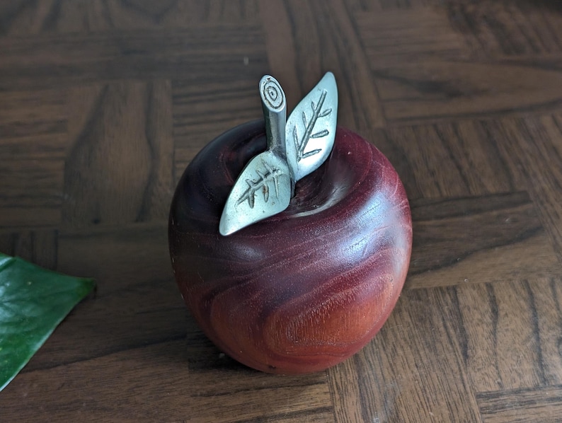 K&ouml;nnte beinhalten: Dekorative Apfelfigur aus rotbraunem Holz mit silberfarbenen Metallblatt- und Stieldetails. Der Apfel hat eine glatte, polierte Oberfl&auml;che. Die Figur ist als dekoratives Objekt gestaltet und soll einem echten Apfel &auml;hneln.