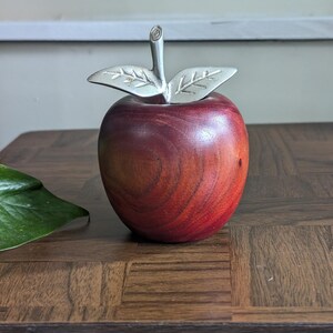 K&ouml;nnte beinhalten: Ein dekorativer Apfel aus Holz mit polierter, rotbrauner Oberfl&auml;che. Er hat einen silberfarbenen Metallstiel und Bl&auml;tter. Der Apfel steht auf einer dunklen Holzoberfl&auml;che, geeignet f&uuml;r die Inneneinrichtung.