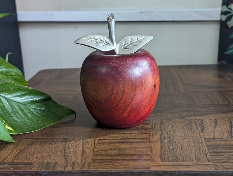 K&ouml;nnte beinhalten: Ein dekorativer Apfel aus Holz mit polierter, rotbrauner Oberfl&auml;che. Er hat einen silberfarbenen Metallstiel und Bl&auml;tter. Der Apfel steht auf einer dunklen Holzoberfl&auml;che, geeignet f&uuml;r die Inneneinrichtung.