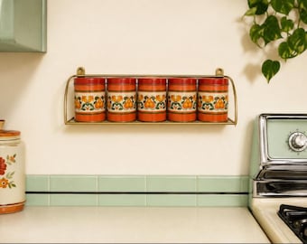 May include: A wall-mounted gold-toned metal rack holding five cylindrical orange canisters. Each canister features a repeating pattern of stylized orange fruits and green leaves with white accents, arranged in a horizontal row on the shelf.