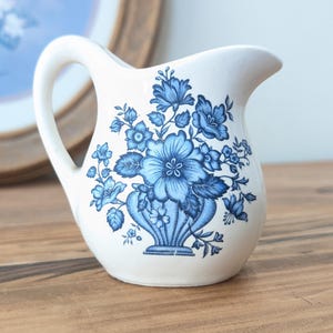May include: A white ceramic milk jug with a blue floral design. The jug has a rounded body and a spout. The floral design features blue flowers and leaves on a white background.
