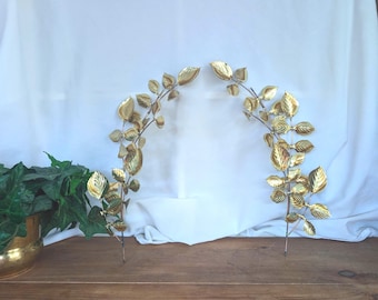 Pode incluir: Dois ramos decorativos de parede feitos de metal polido em tom dourado, cada um com múltiplas folhas texturizadas dispostas ao longo de um caule curvo. Os ramos estão posicionados simetricamente para formar um arco com brilho metálico.