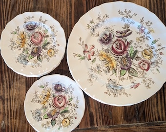 May include: A set of white ceramic plates with scalloped edges, featuring a central floral bouquet illustration in shades of pink, yellow, purple, and green. The collection includes one large dinner plate and two smaller side plates.