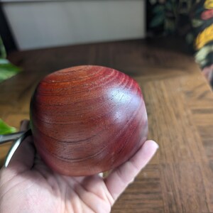 K&ouml;nnte beinhalten: Eine runde, rotbraune Holzschale mit glatter, polierter Oberfl&auml;che. Die Schale wird in einer Hand gehalten, wodurch ihre Gr&ouml;&szlig;e und Textur hervorgehoben werden. Die Holzmaserung ist sichtbar und tr&auml;gt zu ihrer nat&uuml;rlichen &Auml;sthetik bei. Die Schale ist wahrscheinlich f&uuml;r dekorative oder funktionale Zwecke bestimmt.