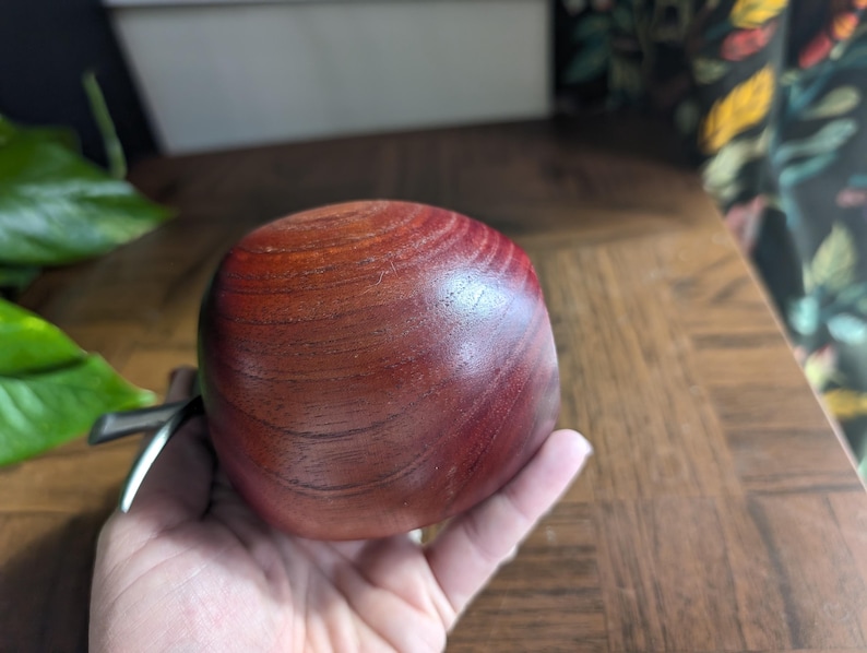 K&ouml;nnte beinhalten: Eine runde, rotbraune Holzschale mit glatter, polierter Oberfl&auml;che. Die Schale wird in einer Hand gehalten, wodurch ihre Gr&ouml;&szlig;e und Textur hervorgehoben werden. Die Holzmaserung ist sichtbar und tr&auml;gt zu ihrer nat&uuml;rlichen &Auml;sthetik bei. Die Schale ist wahrscheinlich f&uuml;r dekorative oder funktionale Zwecke bestimmt.