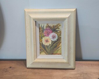 May include: A framed floral painting featuring a bouquet of pink, white, and yellow flowers with green leaves. The artwork is set in a cream-colored, textured frame. The painting is displayed on a wooden surface against a light blue wall.