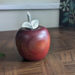 K&ouml;nnte beinhalten: Ein dekorativer Apfel aus Holz mit polierter, rotbrauner Oberfl&auml;che. Er hat einen silberfarbenen Stiel und Bl&auml;tter, die einen Hauch von Eleganz verleihen. Der Apfel ist ein einzigartiger Dekorationsartikel.
