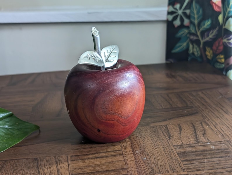 K&ouml;nnte beinhalten: Ein dekorativer Apfel aus Holz mit polierter, rotbrauner Oberfl&auml;che. Er hat einen silberfarbenen Stiel und Bl&auml;tter, die einen Hauch von Eleganz verleihen. Der Apfel ist ein einzigartiger Dekorationsartikel.