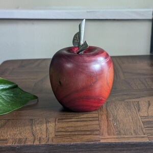 K&ouml;nnte beinhalten: Ein dekorativer Apfel aus Holz mit einem Metallstiel und Blatt. Der Apfel hat eine tiefrote Farbe mit sichtbarer Holzmaserung. Er steht auf einem dunkelbraunen Holztisch. Der Apfel ist ein dekorativer Gegenstand.