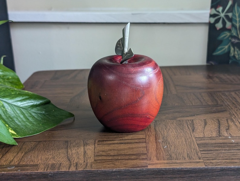 K&ouml;nnte beinhalten: Ein dekorativer Apfel aus Holz mit einem Metallstiel und Blatt. Der Apfel hat eine tiefrote Farbe mit sichtbarer Holzmaserung. Er steht auf einem dunkelbraunen Holztisch. Der Apfel ist ein dekorativer Gegenstand.