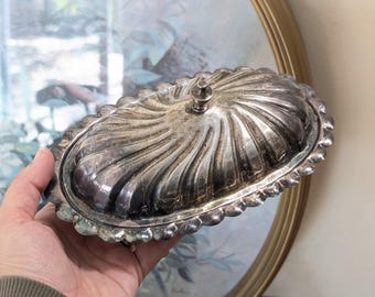 May include: An antique silver-plated butter dish with a lid. The oval dish features a fluted design and a decorative handle on the lid. The edges of the dish have a scalloped trim. The dish is held in a person's hand.