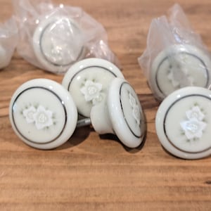 May include: Six white ceramic cabinet knobs with a silver trim and a floral design. The knobs are packaged in plastic bags.