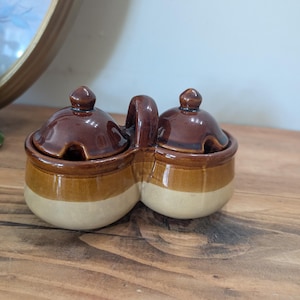 Puede incluir: Un servidor de condimentos de cerámica vintage con dos compartimentos conectados. Cada compartimento tiene una tapa marrón con un pequeño pomo. El servidor es marrón, beige y crema, con un asa entre los compartimentos.